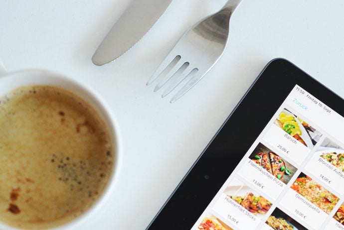 Eine Kaffeetasse, Besteck und ein iPad mit einem Menü liegen auf einem weißen Hintergrund